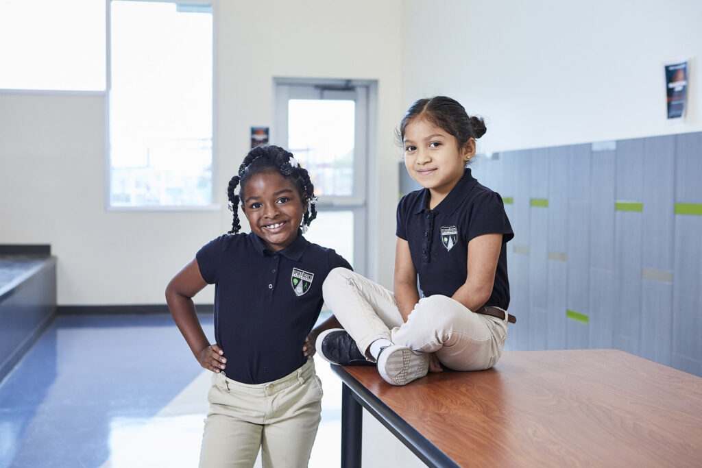 Two School Girls, One Standing And One Sitting, Smiling At The Camera